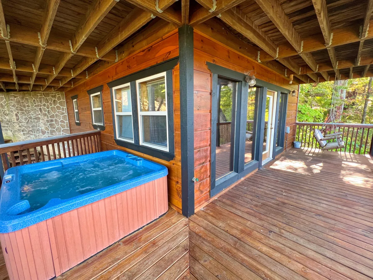 An exterior view of the lower deck featuring a bubbling hot tub against natural surroundings. The deck also shows outdoor seating, and the proximity to windows hints at direct access from the interior game room. The scene is tranquil and inviting, illustrating the indoor-outdoor connection.