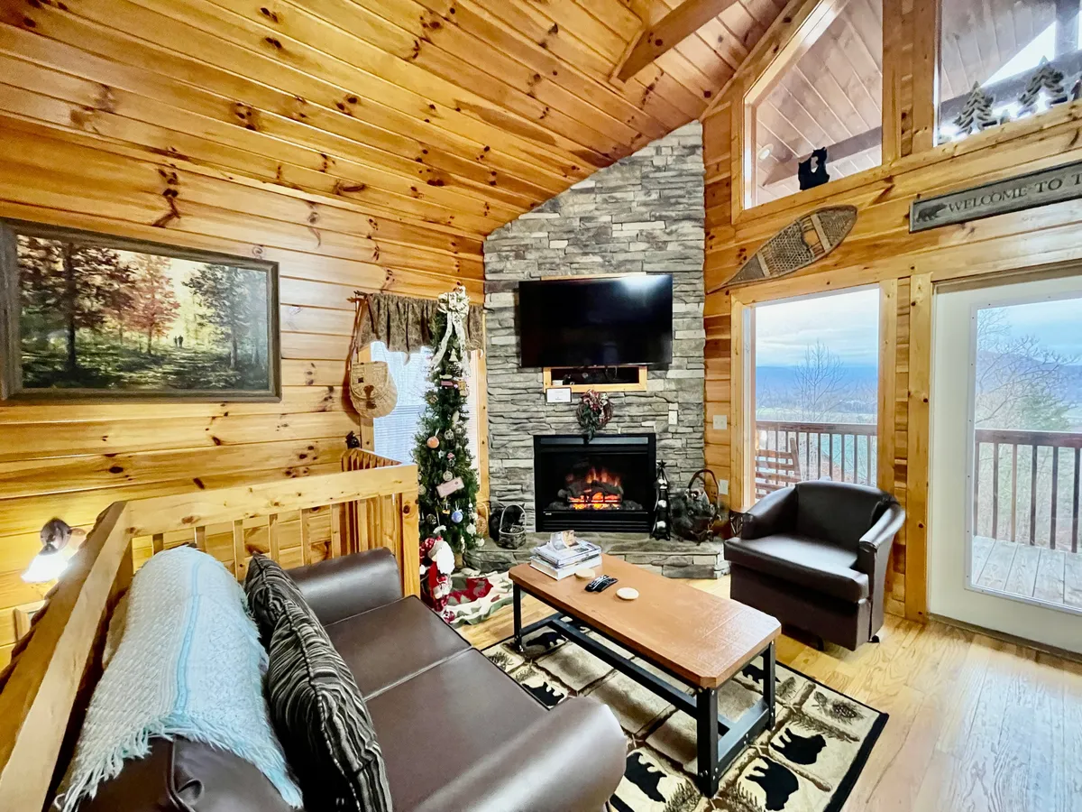 This image captures a warmly lit living room featuring a prominent stone fireplace, a TV mounted above, plush seating, a decorative tree, and exposed wooden beams. The space feels both rustic and inviting—perfect for relaxing with loved ones or enjoying a quiet evening by the fire.