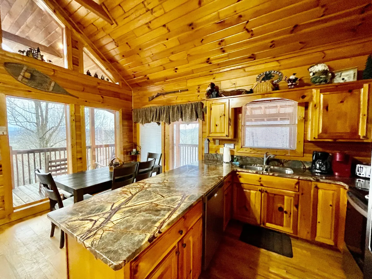 This image presents a warm, welcoming kitchen scene featuring a kitchen island, wooden cabinets, a dining table, and large windows letting in abundant natural light. The open layout visually connects the kitchen and dining area, highlighting both the cooking and gathering spaces.