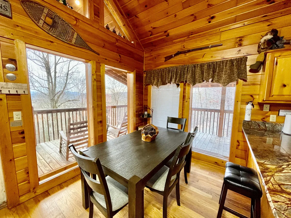 An inviting dining area inside the cabin with a solid wooden table, surrounded by large windows that flood the space with natural light and offer glimpses of the surrounding landscape. The rustic décor and cozy atmosphere evoke the warmth of returning home after a day outdoors.