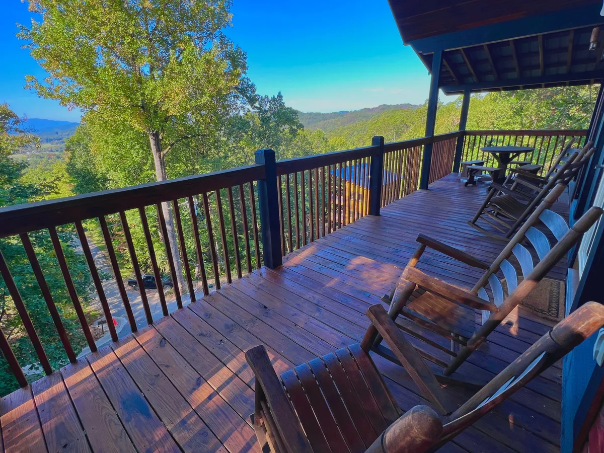 A spacious wooden deck with comfortable rocking chairs facing lush green trees and a distant mountain view. The space is bathed in natural light, inviting guests to sit, relax, and enjoy the serenity of the outdoors.