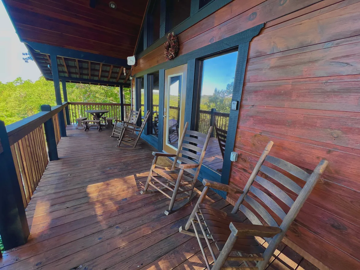 This exterior shot features a welcoming covered deck equipped with classic rocking chairs, a wooden table, sturdy railings, and large windows. The vantage point overlooks tranquil mountain scenery, providing a direct visual connection to the outdoors and the natural beauty of the Smoky Mountains.