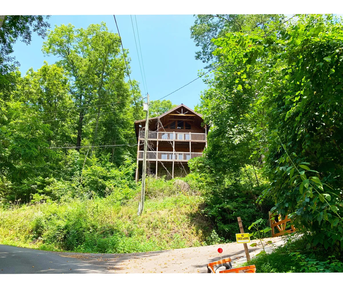 This image shows the wooden exterior of the cabin surrounded by lush greenery, with a balcony visible and a road leading up to the property. The scene evokes a sense of arrival and anticipation for outdoor adventure, highlighting the cabin's proximity to nature and fun family activities.