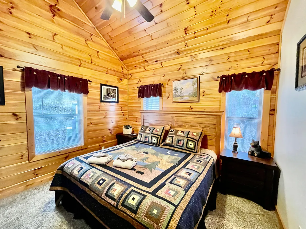 A warmly lit bedroom featuring a king-sized bed with a cozy quilt and wood-paneled walls. Light filters in through curtained windows, and a nightstand with a lamp offers a soft, inviting glow. Framed artwork and rustic accents create a restful, homey atmosphere ideal for relaxing after a busy day.