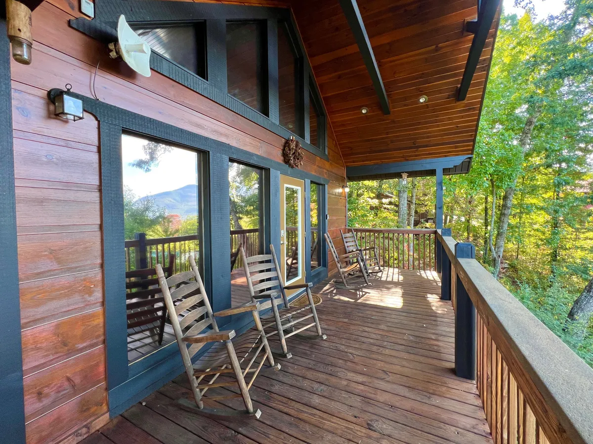 A spacious wooden deck nestled against natural surroundings, featuring a row of classic wooden rocking chairs overlooking scenic mountain views. Large windows reflect the outdoor beauty, creating a welcoming area for relaxation or group gatherings.