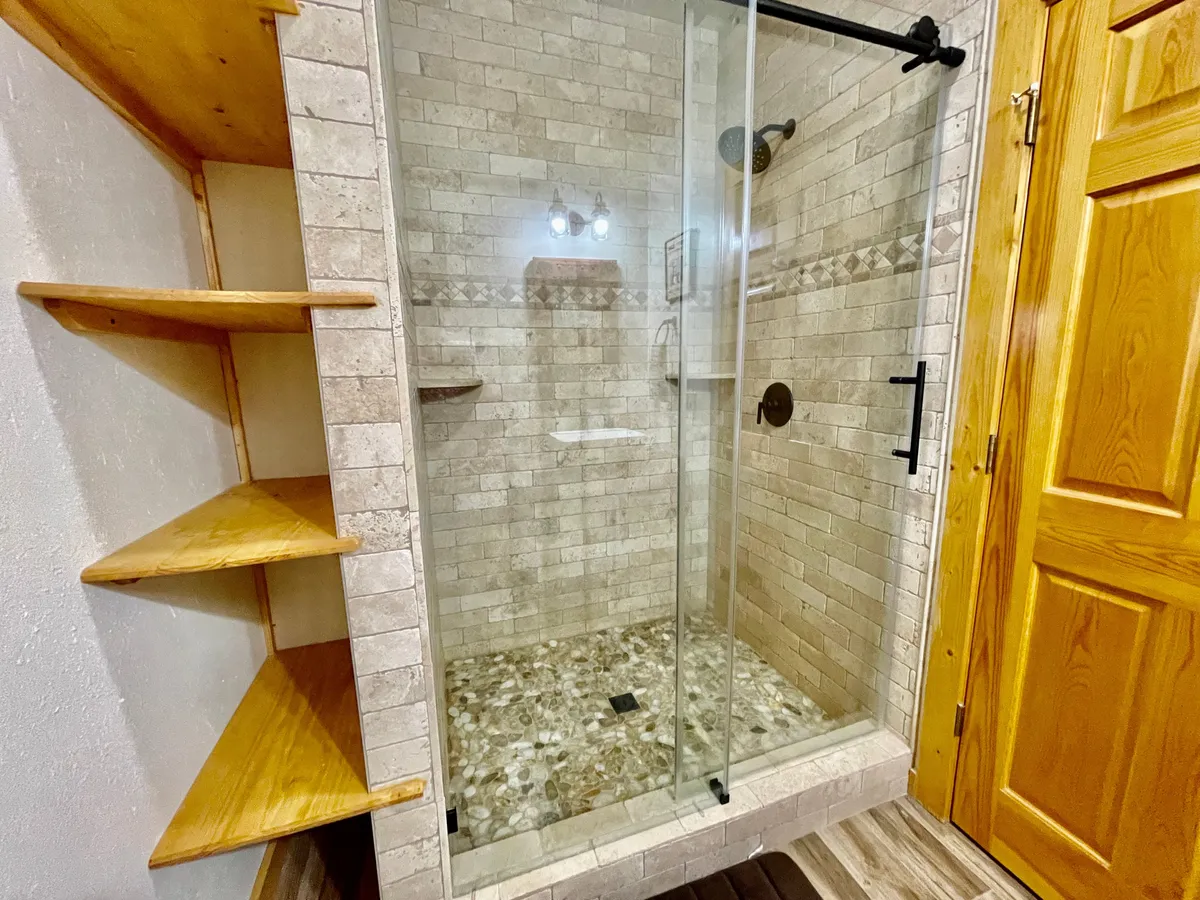 A modern, spa-inspired ensuite bathroom with a glass walk-in shower enclosure, textured stone tiles, and wooden shelving. Sleek metal fixtures and natural materials combine to create a refreshing, serene environment for self-care.