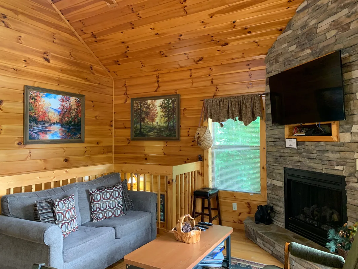 A cozy living area with a soft sofa adorned with patterned cushions, a glowing fireplace, tasteful wall art, and wooden accents. Sunlight streams in through large windows, bathing the room in a warm, welcoming glow.