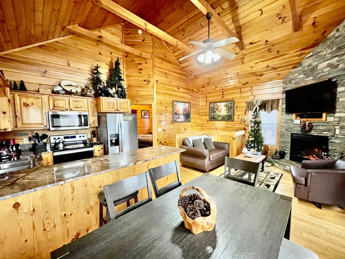 This open-plan space features a wooden ceiling, a large stone fireplace, a dining table for gatherings, and a view into the kitchen area. Decorative touches like pine trees add to the cozy, rustic feel.